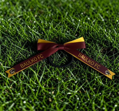 A black hair tie styled into a bow with two maroon and gold ribbon tails printed with 'BAULKO FC' and a small mascot logo, placed on green grass.