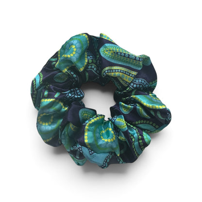 A fabric hair scrunchie featuring a detailed paisley pattern in teal, green, and yellow on a dark navy background, shown on a clean white studio background.