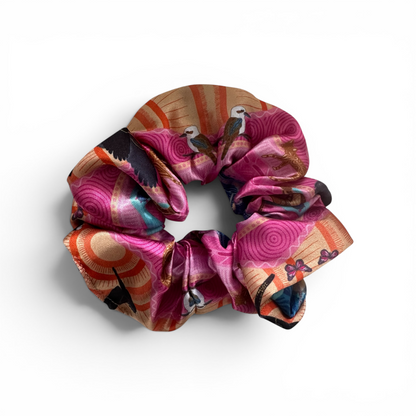 A multicolor silk scrunchie featuring a vibrant pattern of magenta, orange, beige, and black swirls, sun rays, and stylized birds, shown on a clean white background.