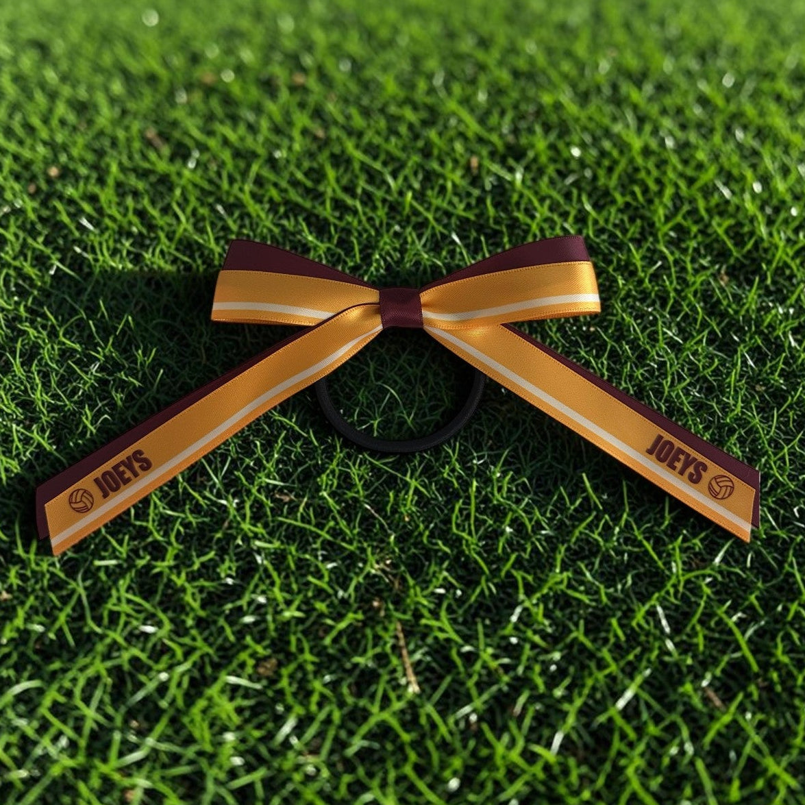 A hair tie bow accessory with gold and maroon ribbon tails, printed with the text 'JOFTS', resting on green grass.