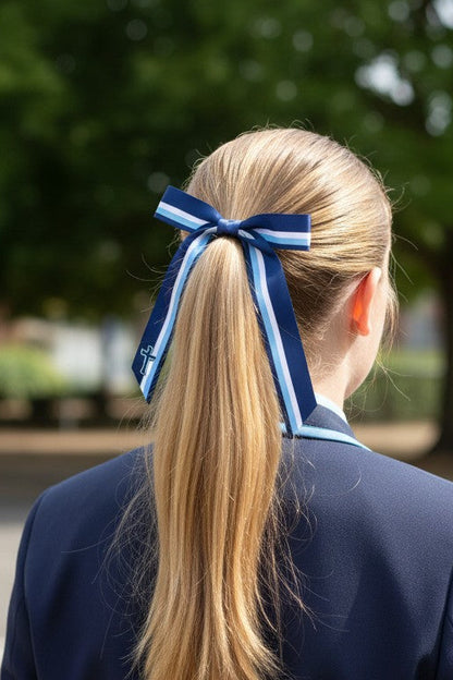 A blonde woman seen from behind wearing a navy blue blazer with light blue side panels and a matching navy and light blue ribbon hair bow in her ponytail, standing outdoors in front of a blurred green tree.