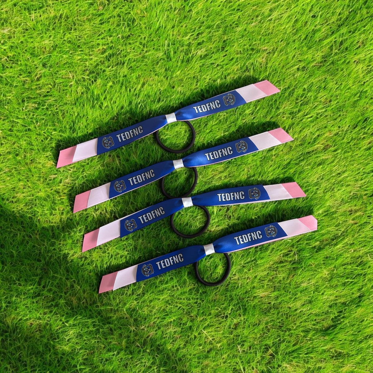 Four blue and pink TED-FNC wristbands with black elastic loops, laid out diagonally on a green grass background.
