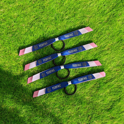 Four blue and pink TED-FNC wristbands with black elastic loops, laid out diagonally on a green grass background.